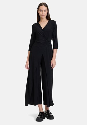 Zwarte jumpsuit van zacht stof, met een wikkeldesign, 3/4 mouwen en wijde benen met een zijsplit. Schoeisel bestaat uit glanzende zwarte loafers.
