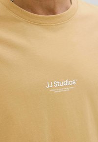 T-shirt jaune moutarde avec un col rond, en coton. Présente un logo "JJ Studios" blanc sur la poitrine et une texture lisse.
