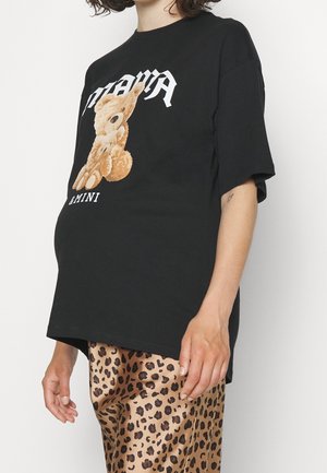 Osoba nosząca czarny, oversize'owy t-shirt z grafiką misia i spodnie w panterkę, stojąca na jednolitym tle.