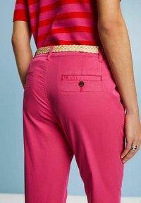 Personne portant un pantalon rose vif avec des poches arrière boutonnées, une ceinture beige tissée et une chemise rayée rouge et rose sur fond bleu.