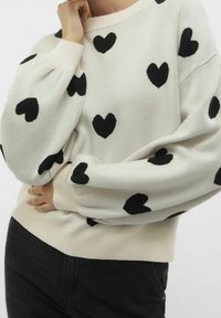 Suéter crema con estampado de corazones negros, con un cuello redondo y puños acanalados. Textura de punto suave, corte relajado y mangas largas.