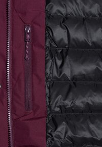 Zbliżenie czarnej pikowanej kurtki z bordowym panelem materiałowym, wyposażonym w pionową kieszeń na zamek błyskawiczny i zęby zamka po lewej stronie.