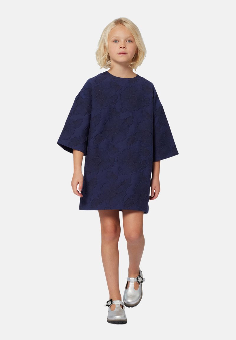 Kind met blond haar in een marineblauwe jurk met bloemenpatroon en zilveren schoenen met zwarte bloemenversieringen, die naar voren loopt.