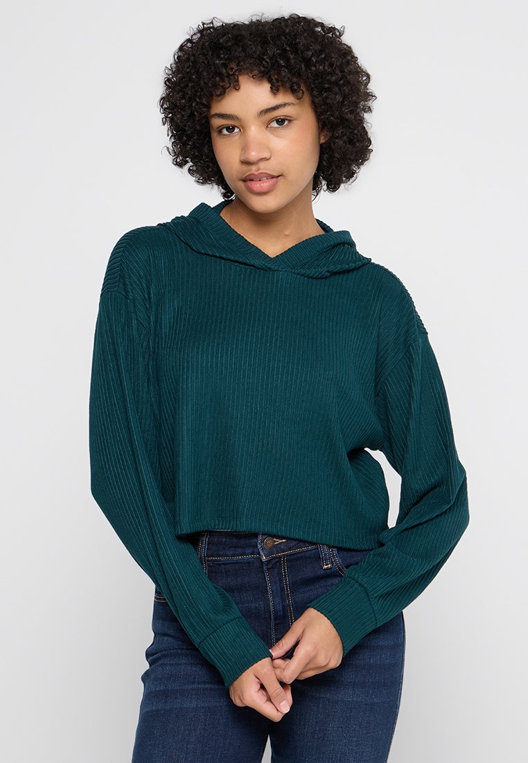 Sudadera corta de color teal con textura acanalada, mangas largas y capucha con cordón, combinada con jeans oscuros. Diseño simple y casual.