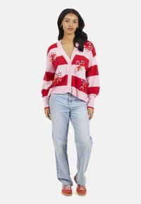 Cardigan a strisce rosse e rosa con ricamo a forma di bastoncino di zucchero, caratterizzato da scollo a V, chiusura con bottoni e polsini a coste, abbinato a jeans azzurri.