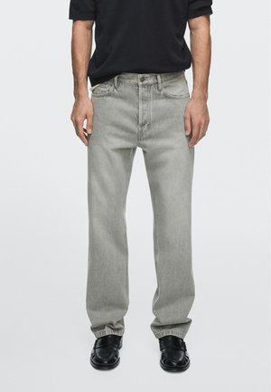 Uomo che indossa jeans dritti grigio chiaro, mocassini neri e una camicia nera, in piedi davanti a uno sfondo semplice.