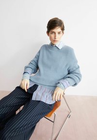 Lichtblauwe gebreide trui over een gestreept overhemd met knopen, gecombineerd met marineblauwe broek met krijtstrepen. Model zittend op een metalen stoel tegen een witte muur.