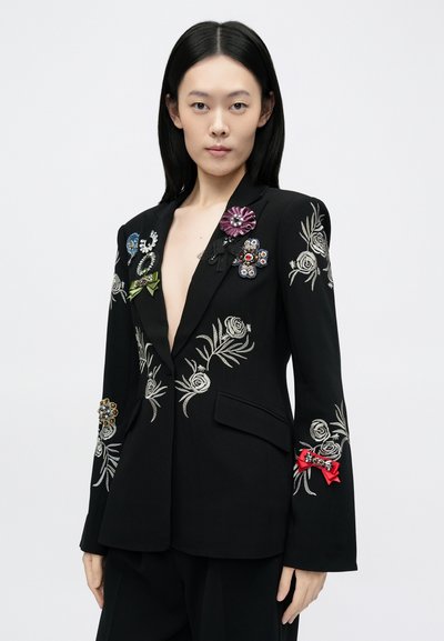 Fekete blézer ezüst virágos hímzéssel, színes kitűzőkkel és szalagokkal díszítve, mély V-nyakkivágással és strukturált sziluettel.
