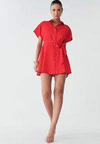 Model in een opvallende rode jurk met een strik in de taille, staande tegen een lichte achtergrond, met minimale accessoires en hakken.
