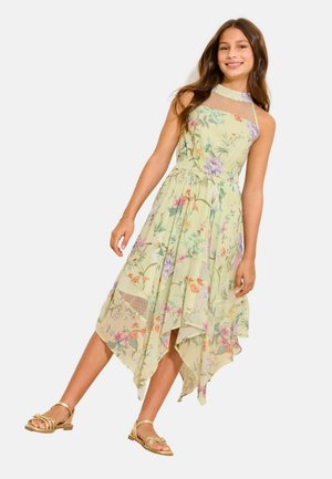 Fille portant une robe sans manches fleurie vert clair avec un ourlet asymétrique et des sandales dorées, souriante et debout sur un fond blanc.