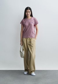 Ροζ T-shirt με κοντό μανίκι σε εφαρμοστή γραμμή με μικρό κεντημένο λογότυπο, συνδυασμένο με μπεζ παντελόνια φαρδιάς γραμμής και λευκά αθλητικά παπούτσια, κρατώντας μια μικρή τσάντα.