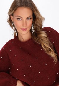Pulover burgundy cu guler rotund, decorat cu pietre strass împrăștiate. Modelul poartă cercei lungi, strălucitori.