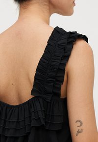 Top nero con tessuto arricciato lungo la spallina e la schiena; presenta una vestibilità ampia e una consistenza morbida. Tatuaggio visibile sul braccio.