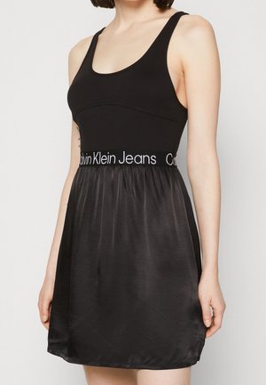 Vestido negro con parte superior tipo tank top ajustada y falda suelta y brillante. Cuenta con una cinturilla elástica con la marca Calvin Klein Jeans. Textura lisa en general.