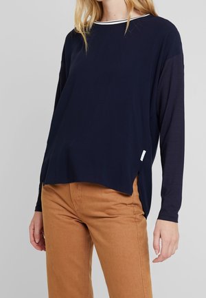 Kobieta ubrana w granatowy top z długim rękawem z białą lamówką przy dekolcie oraz jasnobrązowe spodnie na jednolitym szarym tle.