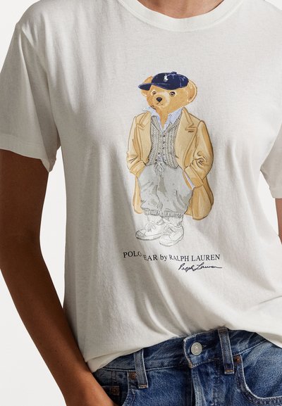 White cotton t-shirt featuring a bear illustration dressed in a beige blazer, gray sweater, and blue cap, with the text "POLO BEAR by RALPH LAUREN."