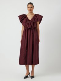 Robe bordeaux avec de grandes manches volantes, décolleté en V, et taille élastique, tombant à mi-mollet. Associée à des escarpins noirs à bout pointu.