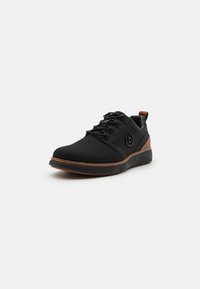 Svart sneakers med en texturerad mesh-övre, bruna accenter vid hälen och sulan, runda snören och en cirkulär logotyp på sidan.