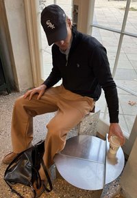 Moški, ki nosi črno kapo in pulover ter bež hlače, sedi na stolčku ob oknu in segá po skodelici kave na okrogli kovinski mizi.