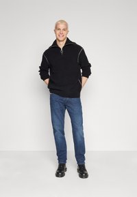 Schwarzer Strickpullover mit hohem Kragen und silbernem Nähdetail, kombiniert mit blauen Jeans und schwarzen Schnürstiefeln.