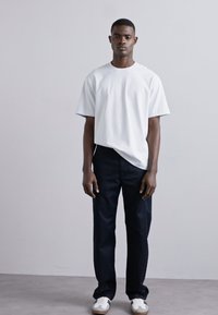 White short-sleeve t-shirt with crew neck, paired with dark navy trousers and white sneakers, standing against a neutral background.