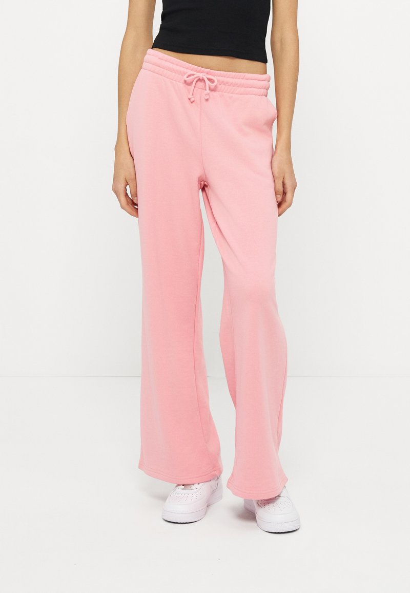 Femme portant un pantalon de survêtement ample rose clair avec une taille à cordon et des baskets blanches, associé à un haut court noir.