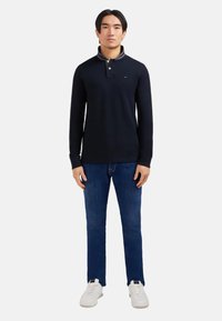 Un homme portant un polo à manches longues bleu marine, un jean bleu et des baskets blanches, se tenant de face contre un fond uni.