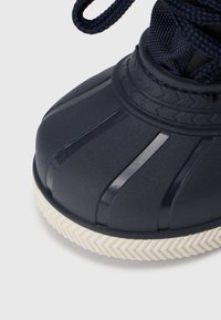 Mørkeblå gummisko med et ribbet design, struktureret tåhætte og kontrasterende hvid zigzag sål. Har snørebåndslukning.