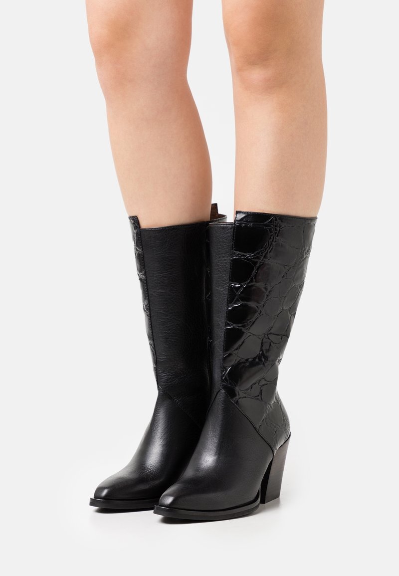 Bottes en cuir noires jusqu'au milieu du mollet avec bouts pointus, talons blocs et motif texturé sur le côté extérieur, portées sur jambes nues.