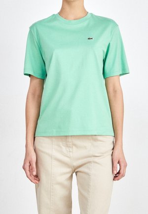 Person trägt ein mintgrünes T-Shirt mit kurzen Ärmeln und einem kleinen Krokodil-Logo sowie beige Hosen und steht vor einem schlichten weißen Hintergrund.