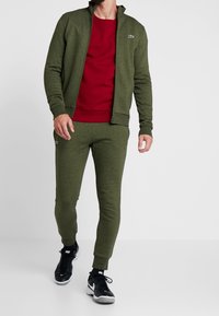 Zelená zipová bunda s vysokým límcem, ladící tepláky a červená mikina. Má přiléhavý střih a elastické manžety. Na nohou jsou černé tenisky.