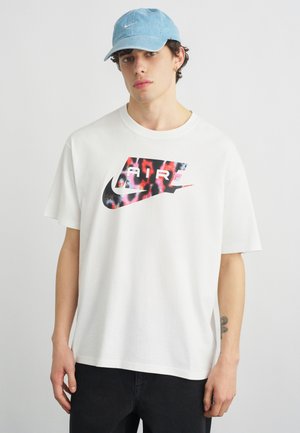 Fiatal férfi világoskék Nike sapkával és fehér, túlméretezett Nike Air pólóval, színes logóval, egyszínű háttér előtt áll.