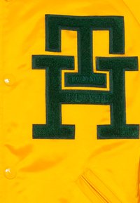 Veste jaune avec un grand logo vert brodé. Le matériau semble lisse avec un éclat. Elle possède des boutons-pression et une poche latérale.