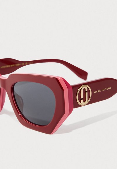 Rode geometrische zonnebril met donkere lenzen en een gouden Marc Jacobs-logo op de arm tegen een witte achtergrond.
