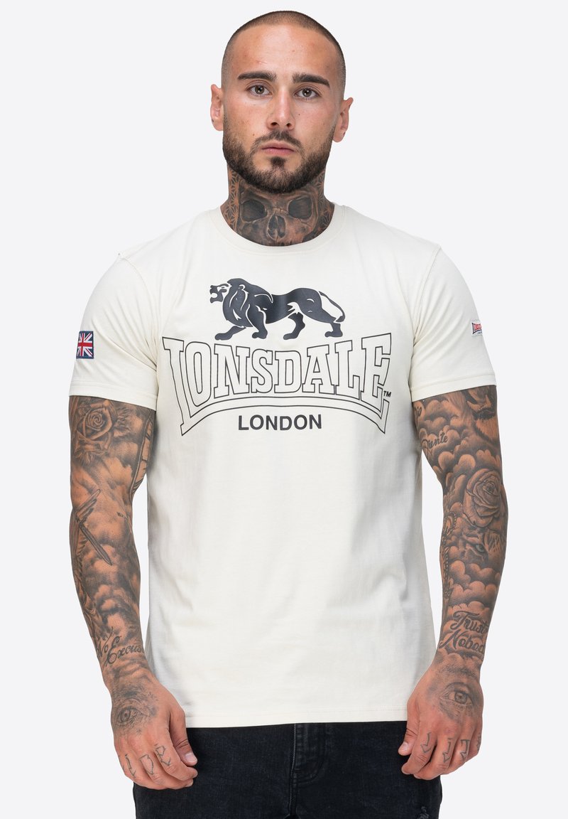 Bijela pamučna majica kratkih rukava s grafikom crnog lava i natpisom "Lonsdale London". Sadrži zakrpu s motivom Union Jack na rukavu.