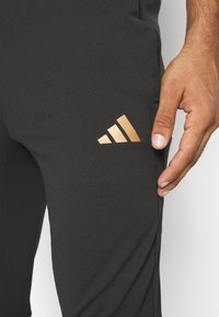 Svarta sportbyxor med en slät yta, försedda med en guldig Adidas-logotyp på låret. Designen inkluderar en åtsittande siluett och en zip-ficka.