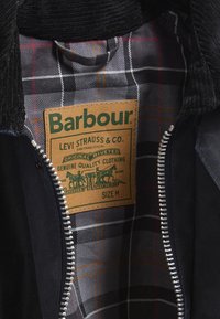 Casaco preto com colarinho de cordura, forro xadrez em cinza e laranja, com um rótulo Barbour cosido na costura interna. Fecho com zíper.