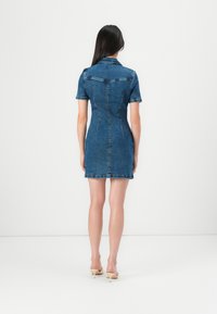 Robe en jean courte avec des manches courtes, forme ajustée et ourlet effiloché. Couleur bleu moyen avec coutures visibles et détail de col.