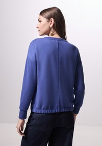 Paarse sweater met een ronde hals, verlaagde schouders, geribbelde manchetten en een gladde textuur. Zichtbare naaddetails langs de achterkant.