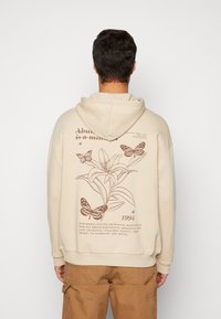 Felpa beige con cappuccio regolabile. Presenta un motivo floreale marrone e farfalle sul retro, insieme a del testo e all'anno 1994.