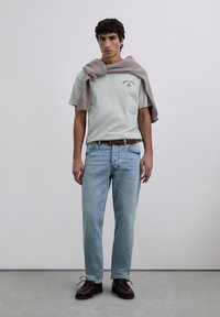 Junger Mann steht da und trägt ein hellgrünes T-Shirt mit Grafik, hellblaue Jeans, einen braunen geflochtenen Gürtel, dunkle braune Schuhe und einen grauen Pullover über den Schultern.