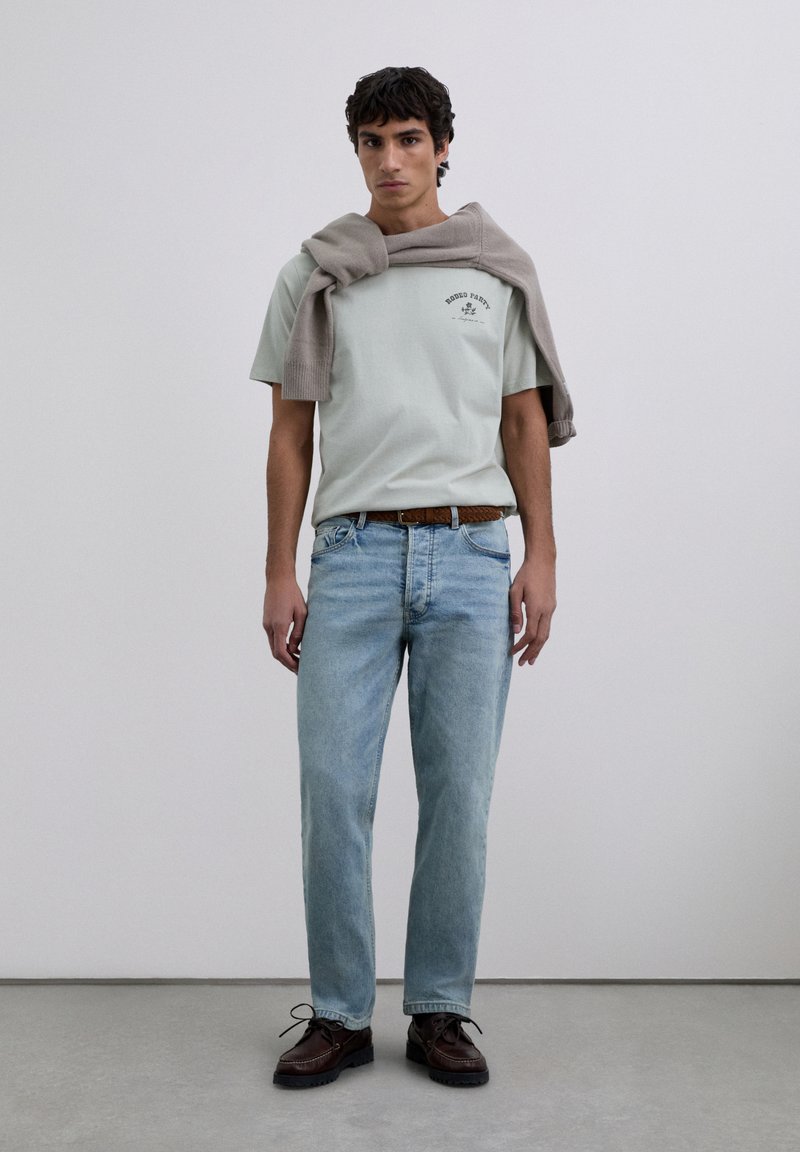 Junger Mann steht da und trägt ein hellgrünes T-Shirt mit Grafik, hellblaue Jeans, einen braunen geflochtenen Gürtel, dunkle braune Schuhe und einen grauen Pullover über den Schultern.