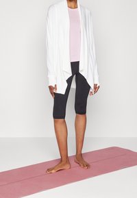Cardigan bianco aperto davanti, top fitted rosa chiaro e leggings neri fino al ginocchio indossati su un tappetino da yoga rosa. La texture appare liscia e leggera.
