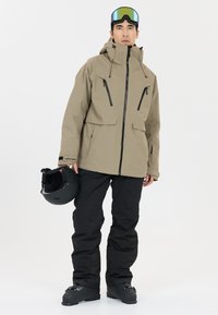 Beige waterdichte jas met zwarte accenten, ritszakken en verstelbare capuchon, gecombineerd met zwarte sneeuwbroeken en een matte zwarte helm.
