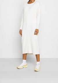 Abito lavorato a maglia di colore crema con texture a coste, scollatura a V, maniche lunghe e lunghezza al polpaccio, abbinato a sneakers sportive bianche e gialle.