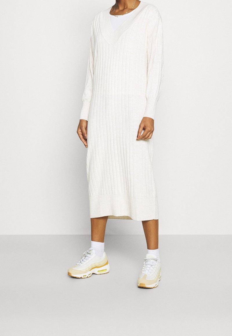 Abito lavorato a maglia di colore crema con texture a coste, scollatura a V, maniche lunghe e lunghezza al polpaccio, abbinato a sneakers sportive bianche e gialle.