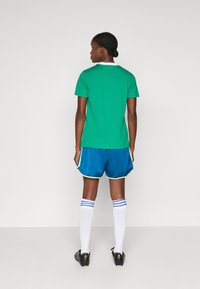 Une personne avec des cheveux tressés se tient de dos, portant un t-shirt vert à manches courtes, un short de sport bleu, des chaussettes blanches jusqu'aux genoux et des chaussures de football noires.