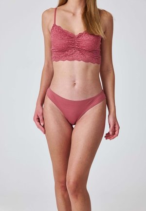 Femme portant un bralette en dentelle rose assorti et un slip uni, debout devant un fond clair uni.