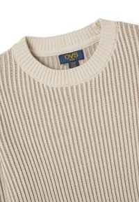Beige rib gebreide trui met een ronde hals. Beschikt over een blauwe label met "OVS kids," maatinformatie en een gemêleerd stofpatroon.