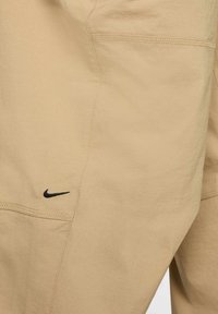 Pantalons Nike beiges en tissu doux avec une texture lisse. Présentent un logo noir et des coutures détaillées le long des bords.